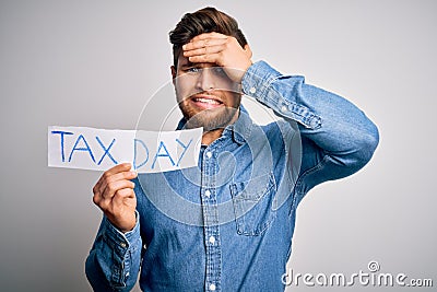Young blond man with beard and blue eyes holding paper with tax day message stressed with hand on head, shocked with shame and Stock Photo