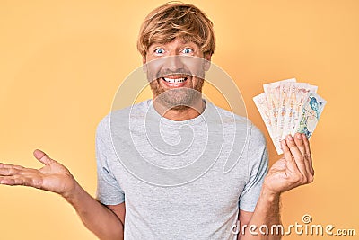 Young blond man holding united kingdom pounds celebrating achievement with happy smile and winner expression with raised hand Editorial Stock Photo