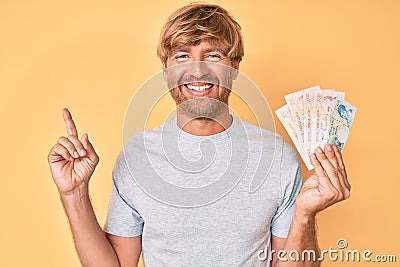 Young blond man holding united kingdom pounds smiling happy pointing with hand and finger to the side Editorial Stock Photo