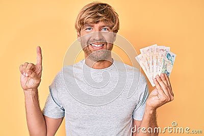 Young blond man holding united kingdom pounds smiling with an idea or question pointing finger with happy face, number one Editorial Stock Photo