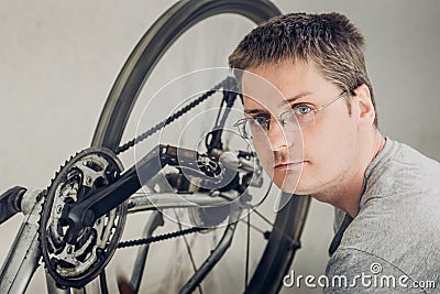 Young blond man with short haircut in a gray t-shirt with glasses sitting near spinning wheel of dirty broken bicycle Stock Photo