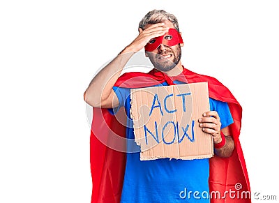 Young blond man wearing super hero costume holding act now cardboard banner stressed and frustrated with hand on head, surprised Stock Photo