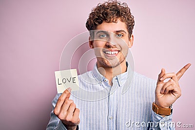Young blond romantic man with curly hair holdiing paper with love message for valentines day very happy pointing with hand and Stock Photo