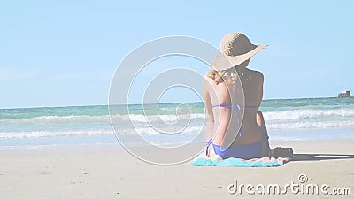 Young blond woman with blue bikini and straw hat sits on the beach and looks at the sea stock video footage Young blond woman with blue bikini and straw hat sits on the beach and looks at the sea stock video footage