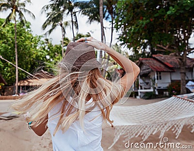 Young blond woman fluttering in hat standing with her back on tropical resort with hammock enjoying vacation Stock Photo