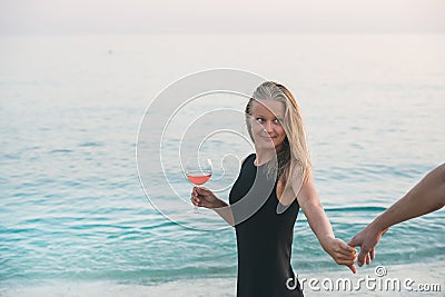 Young blond woman with glass of rose wine holding man's hand on beach by the sea at sunset. Alanya, Turkey. Stock Photo