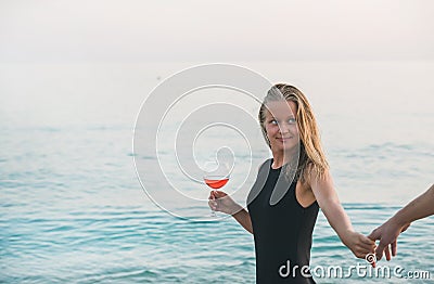 Young blond woman holding glass of rose wine and man's hand on beach by the sea at sunset. Alanya, Turkey. Stock Photo