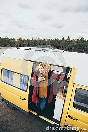 Young blond woman looking out of camper van with solar panel on the roof top and pine forest on the background Stock Photo