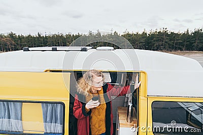 Young blond woman looking out of camper van with solar panel on the roof top and pine forest on the background Stock Photo