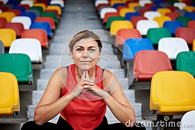 Young blond woman, wearing orange top, black leggings, orange sneakers, sitting on the stairs on stadium tribune, relaxing after Stock Photo