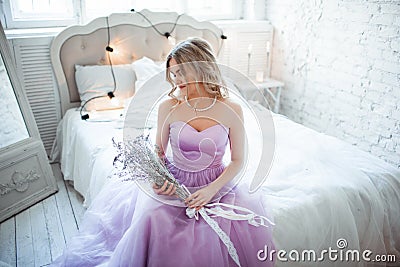 A young blonde bride in a magnificent purple dress sitting on the bed. In the room candles. Girl holds in the hands of Stock Photo