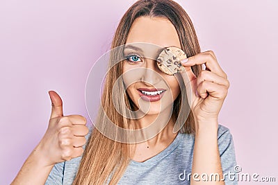 Young blonde girl holding cookie over eye smiling happy and positive, thumb up doing excellent and approval sign Stock Photo
