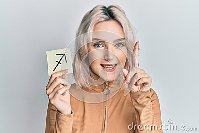 Young blonde girl holding paper with sagittarius zodiac sign smiling with an idea or question pointing finger with happy face, Stock Photo