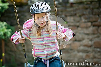 A young blonde girl wearing a cycle helmet while swinging on a garden swing Stock Photo