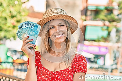 Young blonde tourist woman wearing summer style holding brazilian 100 real banknotes at the city Stock Photo