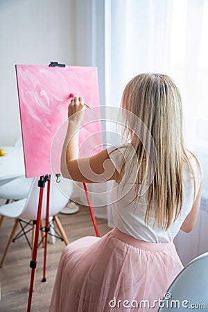 Young blonde woman artist with palette and brush sitting and painting abstract pink picture on canvas near window at Stock Photo