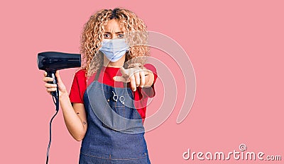 Young blonde woman with curly hair holding dryer blow wearing safety mask for coranvirus pointing with finger to the camera and to Stock Photo