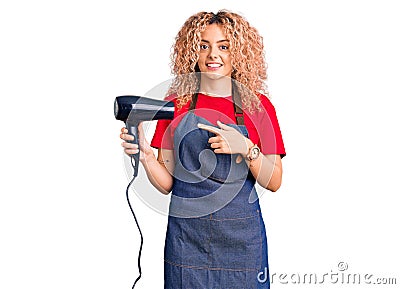 Young blonde woman with curly hair wearing hairdresser apron and holding dryer blow smiling happy pointing with hand and finger Stock Photo