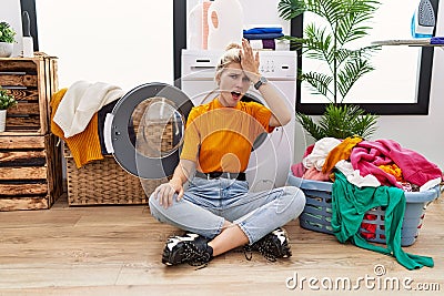 Young blonde woman doing laundry sitting by washing machine surprised with hand on head for mistake, remember error Stock Photo