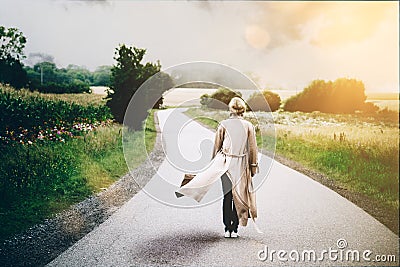 Young blonde woman in flowing long overcoat standing in middle of winding road Stock Photo