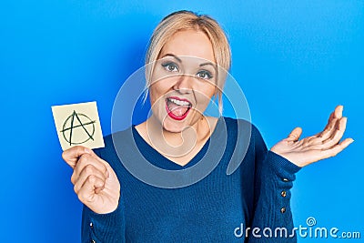 Young blonde woman holding anarchism drawing reminder celebrating achievement with happy smile and winner expression with raised Stock Photo