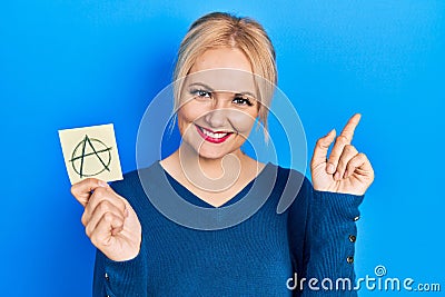 Young blonde woman holding anarchism drawing reminder smiling happy pointing with hand and finger to the side Stock Photo