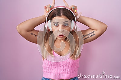 Young blonde woman listening to music using headphones doing funny gesture with finger over head as bull horns Stock Photo