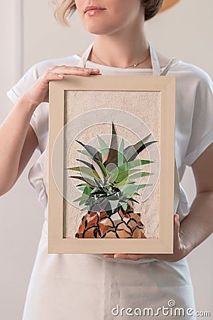 Young blonde woman in a long beige apron is standing in a small workshop and holding in hands mosaic picture in a frame. Stock Photo