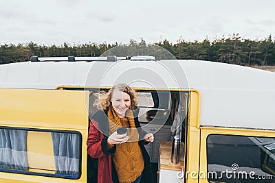 Young blonde woman looking out of camper van with solar panel on the roof top and pine forest on the background Stock Photo
