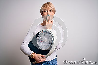 Young blonde woman with short hair holding scale for healthy weight and lifestyle cover mouth with hand shocked with shame for Stock Photo