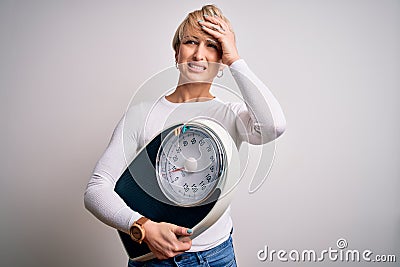 Young blonde woman with short hair holding scale for healthy weight and lifestyle stressed with hand on head, shocked with shame Stock Photo