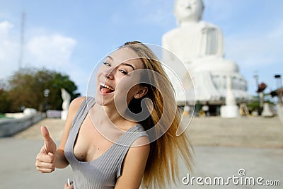 Young blonde woman showing thumbs up, white Buddha statue in Phuket in background. Stock Photo
