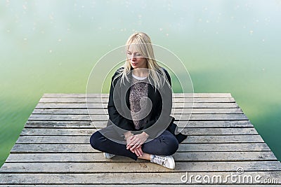 Young blonde woman sitting in pose of lotos on pier in autumn on background of lake Stock Photo