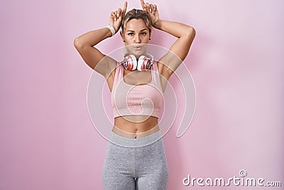 Young blonde woman wearing sportswear and headphones doing funny gesture with finger over head as bull horns Stock Photo