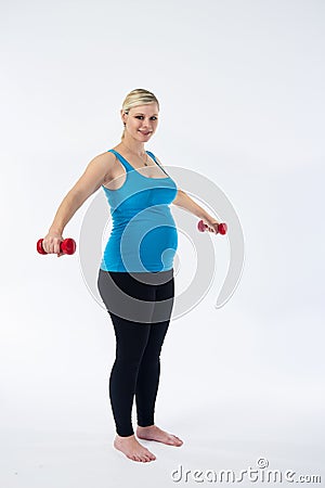 Young blondy pregnant woman in blue tank top does exercises with small red fitness dumbbells, white background. Stock Photo