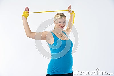 Young blondy pregnant woman in blue tank top does exercises with yellow fitness rubber band, white background. Stock Photo