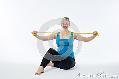 Young blondy pregnant woman in blue tank top does exercises with yellow fitness rubber band, white background. Stock Photo