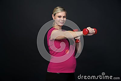 Young blondy pregnant woman in pink t-short with cat does exercises with red fitness dumbbells, black background. Stock Photo