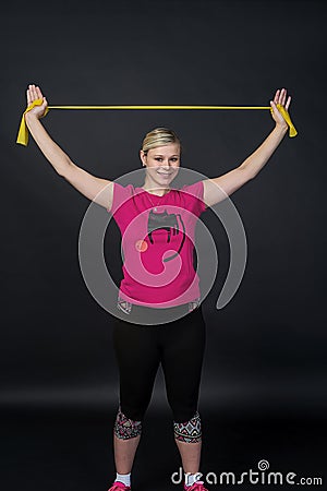 Young blondy pregnant woman in pink t-short with cat does exercises with yellow fitness rubber band, black background. Stock Photo