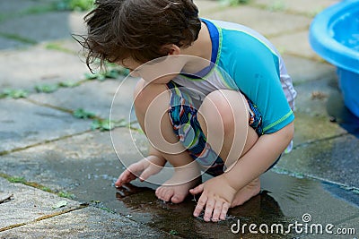 young boy in a bathing suit playing in a puddle of water in the backyard next to a shallow swimming pool Stock Photo