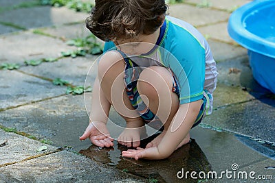 young boy in a bathing suit playing in a puddle of water in the backyard next to a shallow swimming pool Stock Photo