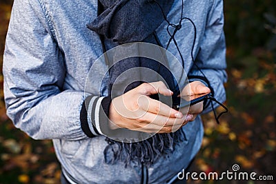 Young boy in gray jacket with gray scarf holds and uses smartphone with headphones outside over autumn background. Technology Stock Photo