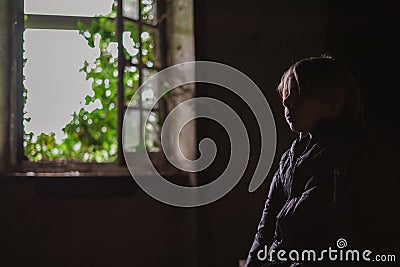 A young boy inside a room in an old crumbling house with an open window. The concept of poverty, homelessness Stock Photo