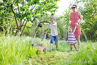 Young boy mowing grass with lawn mower, mother and sister encouraging him Stock Photo