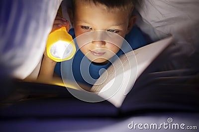 A young boy reading a book under the covers with a flashlight at dark night time Stock Photo