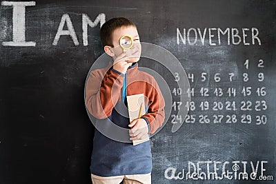 Young boy`s standing with magnifying glass and notebook in his hands near blackboard. Young detective. Creative design Stock Photo