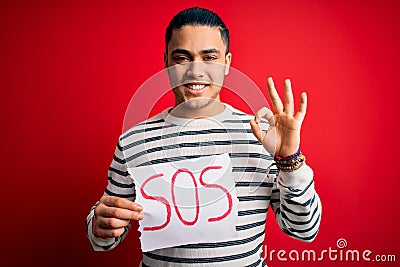 Young brazilian man with problem holding banner with sos message over red background doing ok sign with fingers, excellent symbol Stock Photo