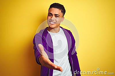 Young brazilian man wearing purple sweatshirt standing over isolated yellow background smiling friendly offering handshake as Stock Photo