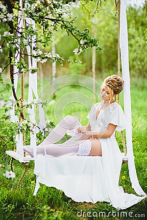 Young bride with blond hair in white negligee and stockings putting on a garter Stock Photo