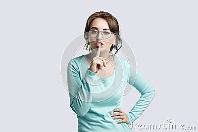 A young brown-haired woman in a mint-colored jumper and in round glasses narrowed her eyes and held her index finger at the lips Stock Photo